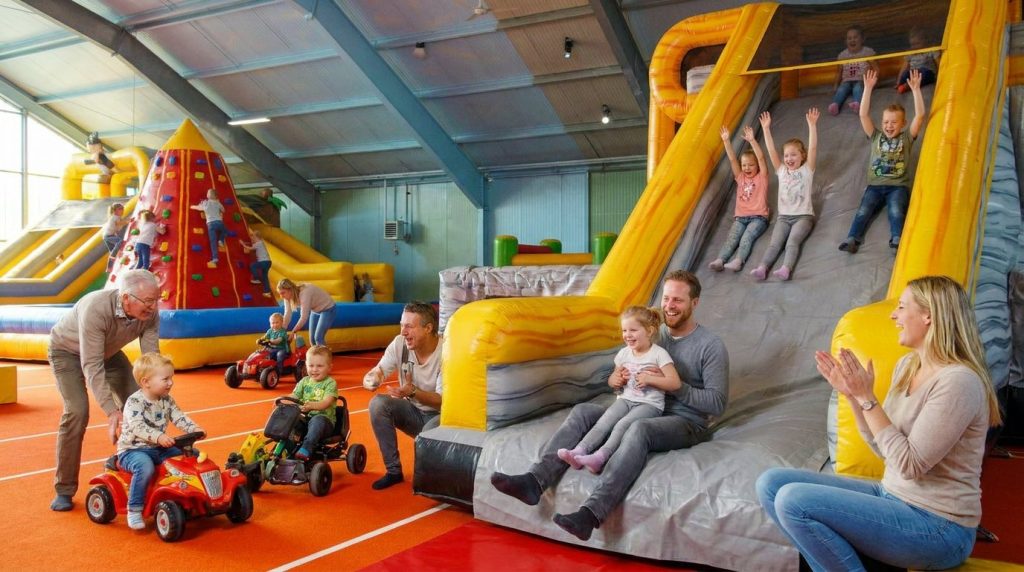 Kinder rutschen jubelnd die große Rutsche im Kinderparadies Landsberg hinunter, unten sitzt ein Vater mit seiner Tochter, Mutter und Großvater freuen sich mit; im Hintergrund Kletterpyramide und Kinder auf Kinderfahrzeugen